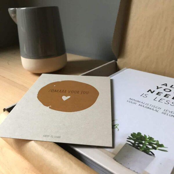 Boek all you need is less met zomaar voor jou kaart in cadeau doosje met kraft bruin zijdepapier op blank houten tafel met minimalistisch kannetje.