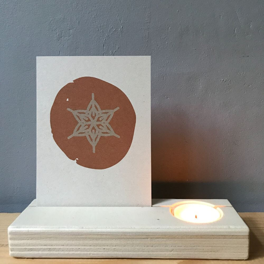 Blank houten kaartenhouder met brandend theelichtje en natuurlijke vezelrijke kerstkaart met een ster in ecru met cognac.