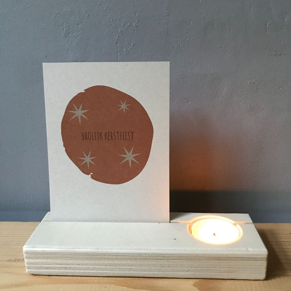 Blank houten kaartenhouder met brandend theelichtje en natuurlijke vezelrijke kerstkaart met de tekst vrolijk kerstfeest in ecru met cognac.
