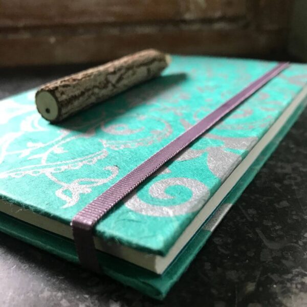 Groen notitieboekje met zilveren bloemversiering en een sluitelastiek met potlood van boomschors.