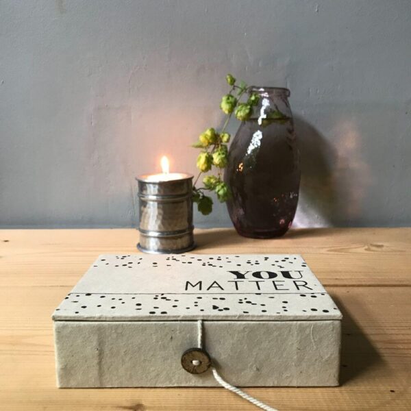 Ecru kleurige you matter box van handgeschept papier houten tafel met brandend kaarsje en bloemenvaasje.